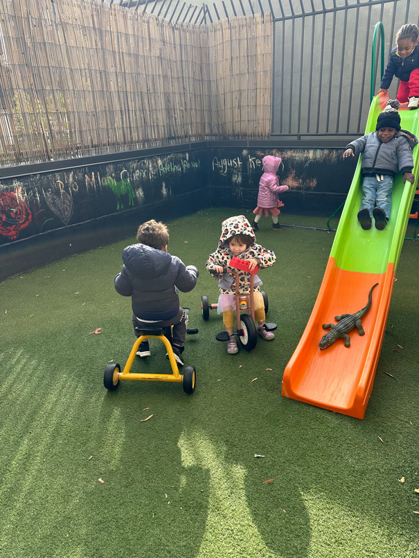 Children playing on slides and tricycles during outdoor rooftop time at Two By Two